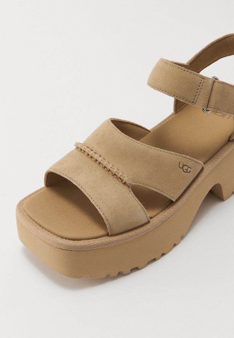 Beige Wildledersandalen mit zwei breiten Riemen, verstellbarem Knöchelriemen, dicker Plattformsohle und subtiler UGG-Branding an der Seite.
