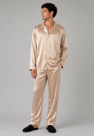 Homme portant un haut de pyjama en satin beige à manches longues boutonné et un pantalon assorti avec des chaussons noirs, debout devant un fond gris uni.