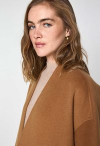 Cardigan avvolgente in misto lana color beige chiaro con una texture morbida e una vestibilità rilassata, dotato di scollatura a V e cuciture minime. Completato da una maglietta beige chiaro.