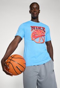 Blå Nike T-shirt med et rødt basketballgrafik, parret med grå sportsbukser. Modellen holder en orange basketball.