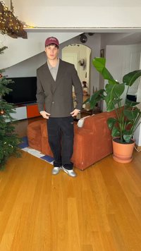 Σκούρο καφέ διπλόμπερτο σακάκι πάνω από γκρι πόλο, μαύρο παντελόνι και ασημένια αθλητικά παπούτσια, όρθιος σε δωμάτιο με φυτά και ξύλινο πάτωμα.