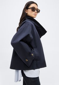 Γυναίκα που φοράει υπερμεγέθη ναυτικό μπουφάν με ψηλό γιακά, γυαλιά ηλίου από καφέ διαφανές πλαστικό, λευκό πουκάμισο και σκούρο τζιν, πόζα πλαγίως σε λευκό φόντο.