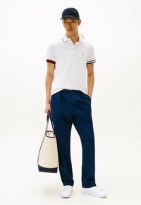 Witte polo met zwarte en rode streepaccenten, marineblauwe broek, witte sneakers en een marineblauwe pet, houdend een crèmekleurige tas met marineblauwe afwerking.