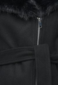 Čierny kabát s umelým kožušinovým golierom, má strieborný zips a opasok na viazanie. Materiál vyzerá jemne a má štruktúrovaný povrch.