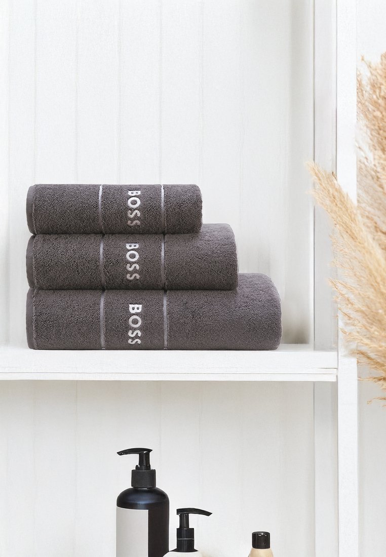 En stapel av tre mörkgrå handdukar med "BOSS" broderat i vitt, med en mjuk textur och rena linjer, placerade på en vit hylla.