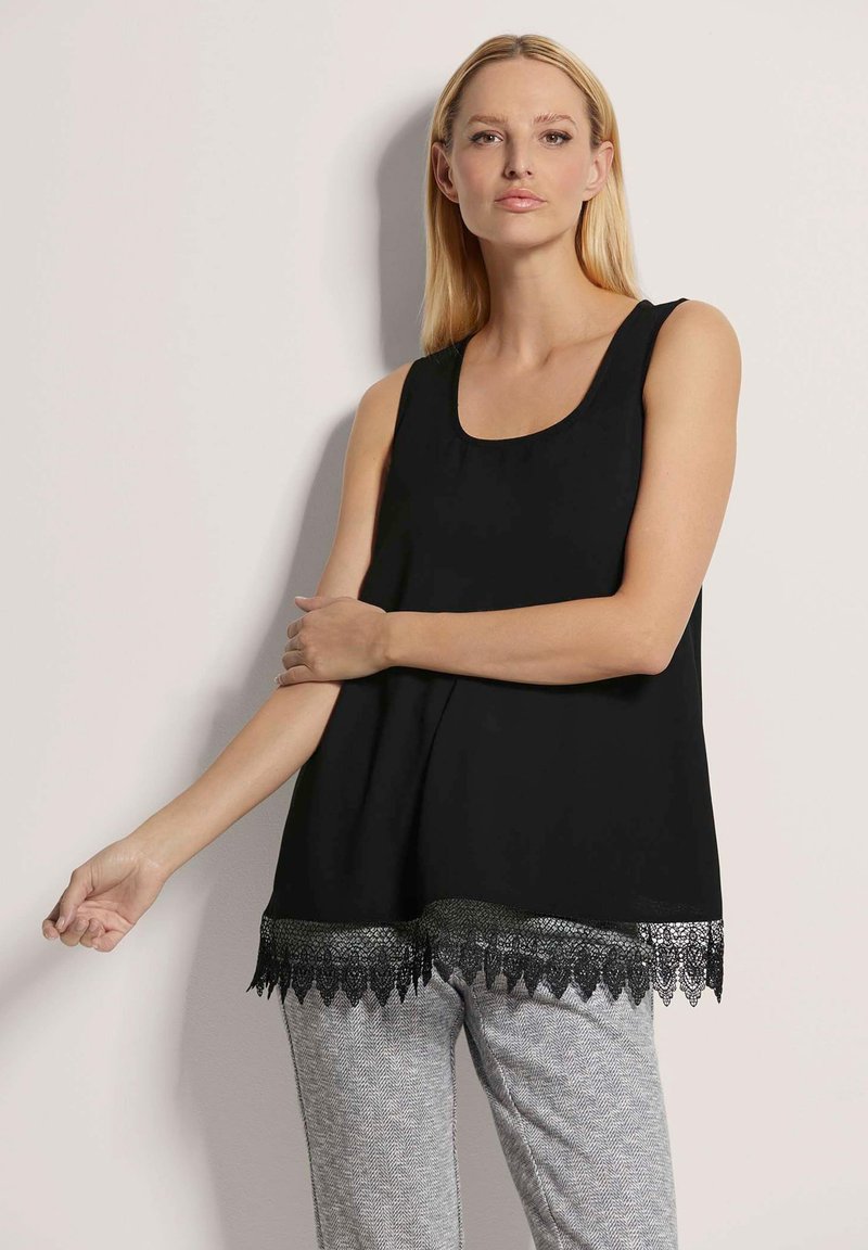 Schwarzes ärmelloses Oberteil mit einem runden Ausschnitt und einer Spitzenborte, die ein gewelltes Muster am Saum aufweist. Kombiniert mit hellgrauen gemusterten Hosen.