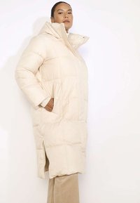 Bēšas krāsas pufīgs garais mēteli ar augstu apkakli, sānu kabatām un priekšējo rāvējslēdzēju. Iezīmē horizontālu kviltētu rakstu un sānu šķēlumu.