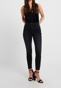 Svarta högmidjade skinny jeans med en slät yta, med fem fickor och knappstängning, i kombination med en svart plisserad topp och klackar med remmar.