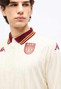 Νεαρός άνδρας που φοράει κρεμ αθλητική φανέλα με γιακά σε μαρορινή και χρυσή απόχρωση, λογότυπο ομάδας με αστέρια στο στήθος και λογότυπα μάρκας στους ώμους.