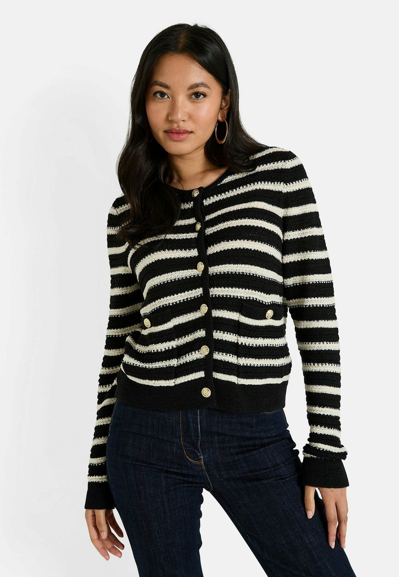 Femme aux longs cheveux noirs, portant des boucles d'oreilles en or, un cardigan à rayures noir et blanc boutonné et un jean bleu foncé, sur un fond blanc.