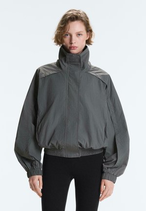 Jonge persoon draagt een grijze, loszittende windjack met hoge kraag, elastische manchetten en zoom, gecombineerd met zwarte smalle broek.