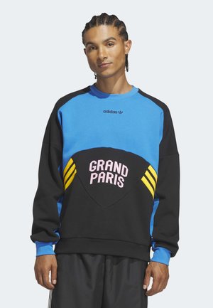 Jeune homme aux cheveux tressés porte un sweat-shirt Adidas bleu et noir avec des rayures jaunes et le texte "GRAND PARIS" sur le devant.
