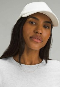 Weißer Hut mit gebogenem Schirm, getragen mit einem hellgrauen T-Shirt. Der Hut hat eine glatte Textur und ein neutrales Design, das eine zarte Kettenhalskette ergänzt.