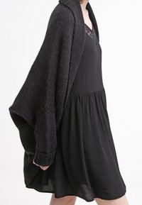 Cardigan de punto negro con diseño oversized, cuello acanalado y mangas largas, llevado sobre un vestido negro sin mangas. Textura suave.