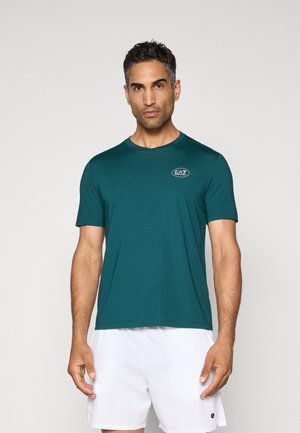 Mann som har på seg en teal EA7-logo t-skjorte og hvite shorts, stående mot en ensfarget lys bakgrunn, vender fremover med et nøytralt uttrykk.