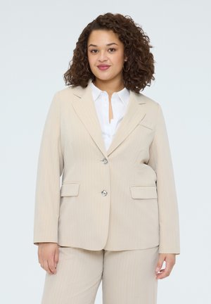 Kiharatukkainen nainen pukeutuu beigenväriseen pukuun, jossa on takki ja samansävyiset housut valkoisen paidan päällä. Hän katsoo eteenpäin ja hymyilee kevyesti.