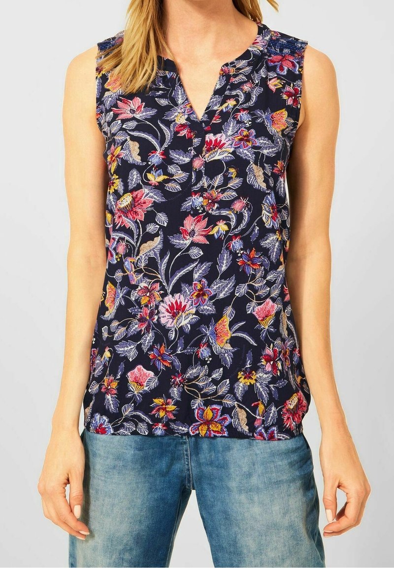 Femme portant un haut sans manches bleu marine avec un motif floral coloré et un col en V, associé à un jean bleu sur fond uni.