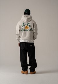 Sivá mikina s kapucňou s textom v zelenej kurzíve na chrbte, v kombinácii s čiernymi voľnými nohavicami a béžovými čižmami, pohľad zozadu.