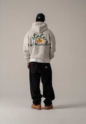 Grijze capuchontrui met groene cursieve tekst op de achterkant, gecombineerd met zwarte loszittende broek en beige laarzen, gezien van achteren.