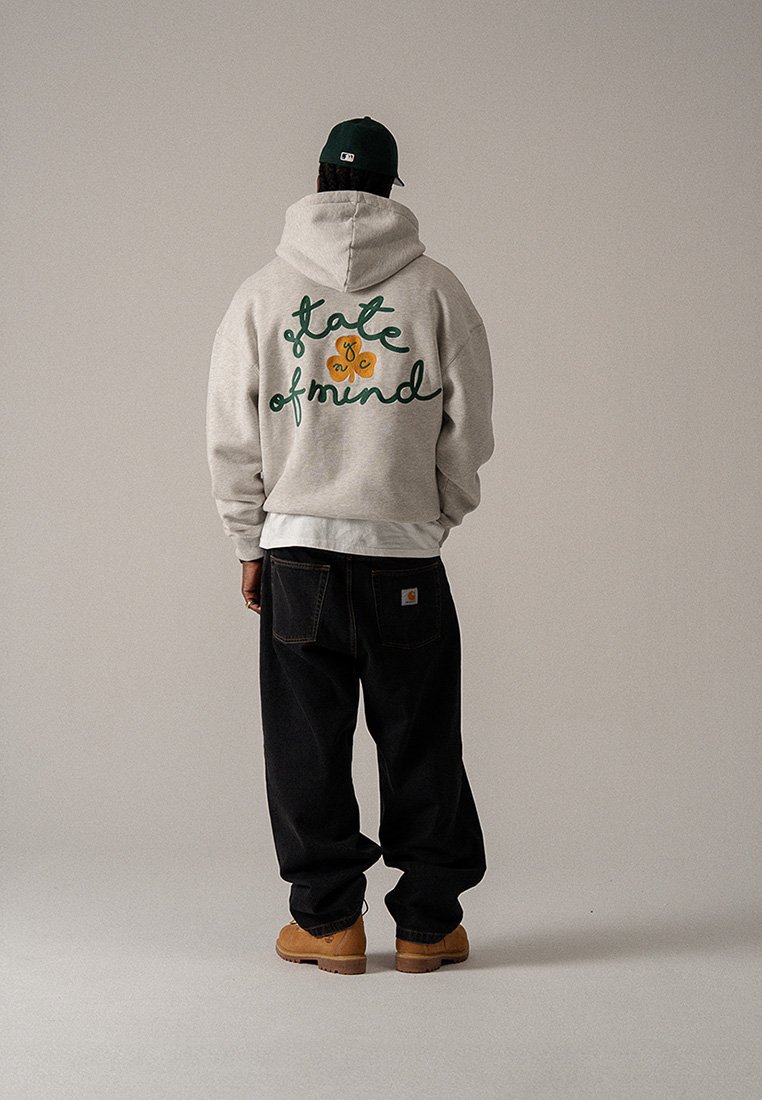 Sivá mikina s kapucňou s textom v zelenej kurzíve na chrbte, v kombinácii s čiernymi voľnými nohavicami a béžovými čižmami, pohľad zozadu.