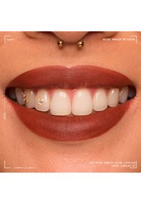 Gros plan d'un sourire avec un rouge à lèvres mat couleur cacao, un piercing septum doré et des strass sur deux dents de devant.
