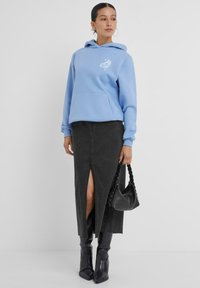 Hoodie azzurro con tasca frontale e design grafico bianco, abbinato a una gonna di jeans nera con spacco laterale e stivali neri hasta al ginocchio.