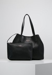 Deux sacs Guess noirs texturés sur une surface blanche, un grand tote avec des sangles pour épaule et une petite pochette zippée devant.