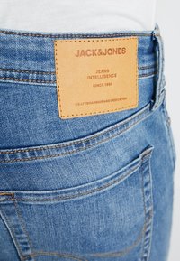 Jeans de mezclilla azul con un parche de cuero marrón en la parte trasera que presenta texto en relieve. Detalles de costura notables y diseño clásico de bolsillo.