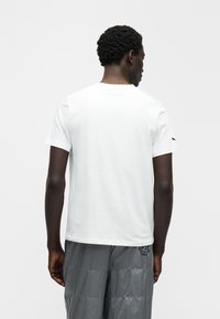 Λευκό t-shirt με κοντά μανίκια, βαμβακερό ύφασμα, στρογγυλή λαιμόκοψη, απαλή υφή, διαθέτει μαύρο λογότυπο στον αριστερό ώμο, μινιμαλιστικός σχεδιασμός.