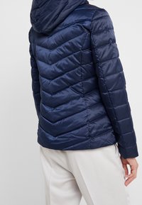 Mörkblå vadderad jacka med fiskbenssömmar, huva och slät, glänsande yta. Har en figurnära passform och elastiska muddar.