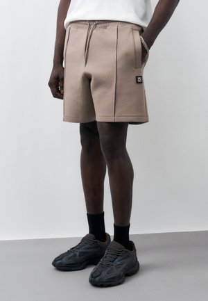 Άνδρας με μπεζ σορτς με κορδόνι στη μέση και μπροστινές τσέπες, μαύρες κάλτσες και μαύρα αθλητικά παπούτσια στέκεται μπροστά σε γκρι και λευκό φόντο.