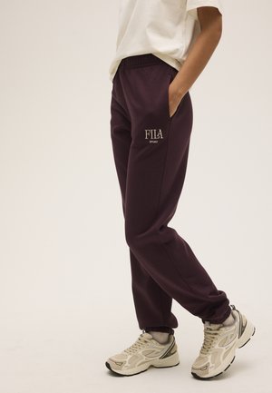 Persona que lleva puestos pantalones deportivos FILA de color púrpura oscuro, camiseta blanca y zapatillas beige, de pie con las manos en los bolsillos.