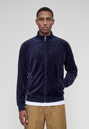 Homme portant une veste zippée en velours bleu marine avec poignets et col côtelés, un t-shirt blanc et un pantalon marron clair sur un fond uni.