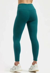 Leggings taille haute couleur sarcelle fabriqués dans un tissu extensible, dotés d'un design sans coutures et d'une texture lisse, associés à des chaussures de sport blanches.