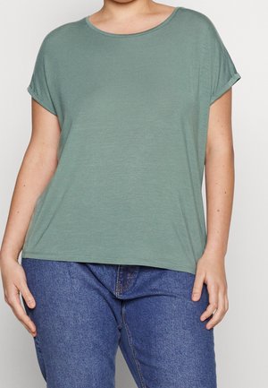 Person wearing a loose-fitting short-sleeve sage green top and blue jeans, standing with relaxed arms against a plain background.