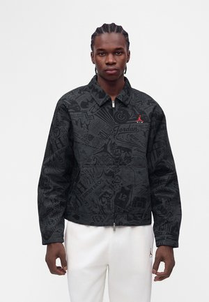 Jonge man met gevlochten haar die een zwarte Jordan-jas met patroon en witte broek draagt, staand tegen een effen lichte achtergrond.