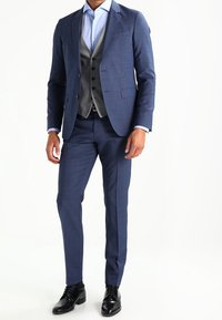 Completo blu scuro con finitura texturizzata, gilet grigio e camicia azzurra; abbinato a scarpe eleganti nere. Fit su misura e design classico.