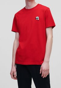 Rød bomulds t-shirt med korte ærmer og rund hals, prydet med et lille sort-hvidt tegneserieansigt på venstre bryst.