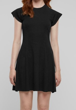 Kvinde iført en kort sort kjole med strukturerede kasketærmer, stående mod en ensartet lys grå baggrund.