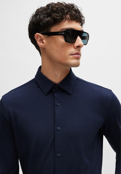 Chemise à manches longues bleu marine avec col et patte de boutonnage, associée à des lunettes de soleil rectangulaires noires avec des verres sombres et des accents métalliques.