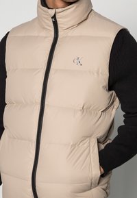 Beige pufferväst med hög krage, frontdragkedja och sidofickor. Visas på en svart tröja och har en liten CK-logotyp på bröstet.