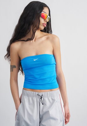 Femme aux longs cheveux foncés portant des lunettes de sport réfléchissantes, un haut Nike bleu bustier et un short gris clair à cordon, les mains dans les poches.