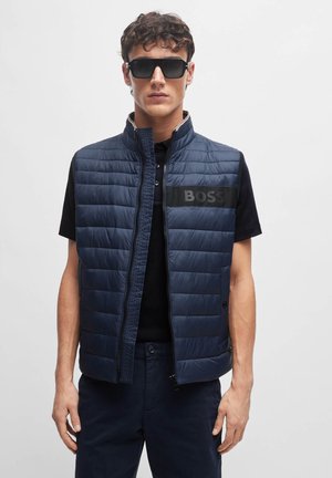 Dunkelblaue, gepolsterte Weste mit hohem Kragen, Front-Reißverschluss und schwarzem Logo-Patch. Kombiniert mit einem schwarzen Shirt und dunkelblauen Hose.