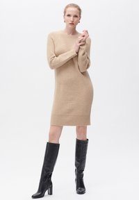 Vestito maglione beige lavorato a maglia con un motivo a spina di pesce testurizzato, polsini a coste e una linea dritta. Abbinato a stivali neri in pelle al ginocchio.