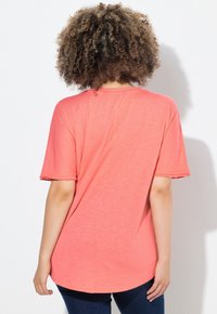 Camiseta coral de corte holgado con mangas cortas enrolladas, dobladillo redondeado y tela suave y texturizada. La vista trasera resalta la simplicidad del diseño.
