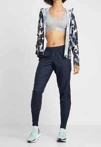 Sportoutfit met een gepatternde zwart-grijze zip-up jack, grijze sportbeha en nauwsluitende marineblauwe broek, gecombineerd met lichtblauwe sneakers.