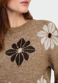 Mujer vistiendo un suéter de punto marrón con grandes patrones de flores en blanco y negro, mostrando el escote y parte de la cara y el cabello.