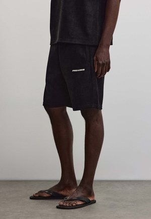 Mann trägt schwarze Cordhosen-Shorts mit „Pegador“-Logo und schwarze Flip-Flops, steht vor einem schlichten grauen Hintergrund.