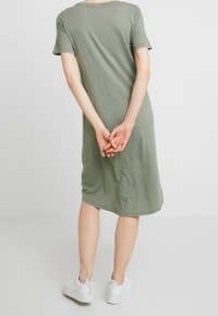 Oliwkowa, krótko rękawowa sukienka T-shirt, długości do kolan, luźny krój, wykonana z miękkiej, teksturowanej tkaniny, noszona z białymi trampkami. Ręce splecione za plecami.