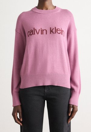 Personne portant un pull rose à manches longues avec le texte "calvin klein" et un pantalon noir, montrée du cou aux hanches sur un fond uni.